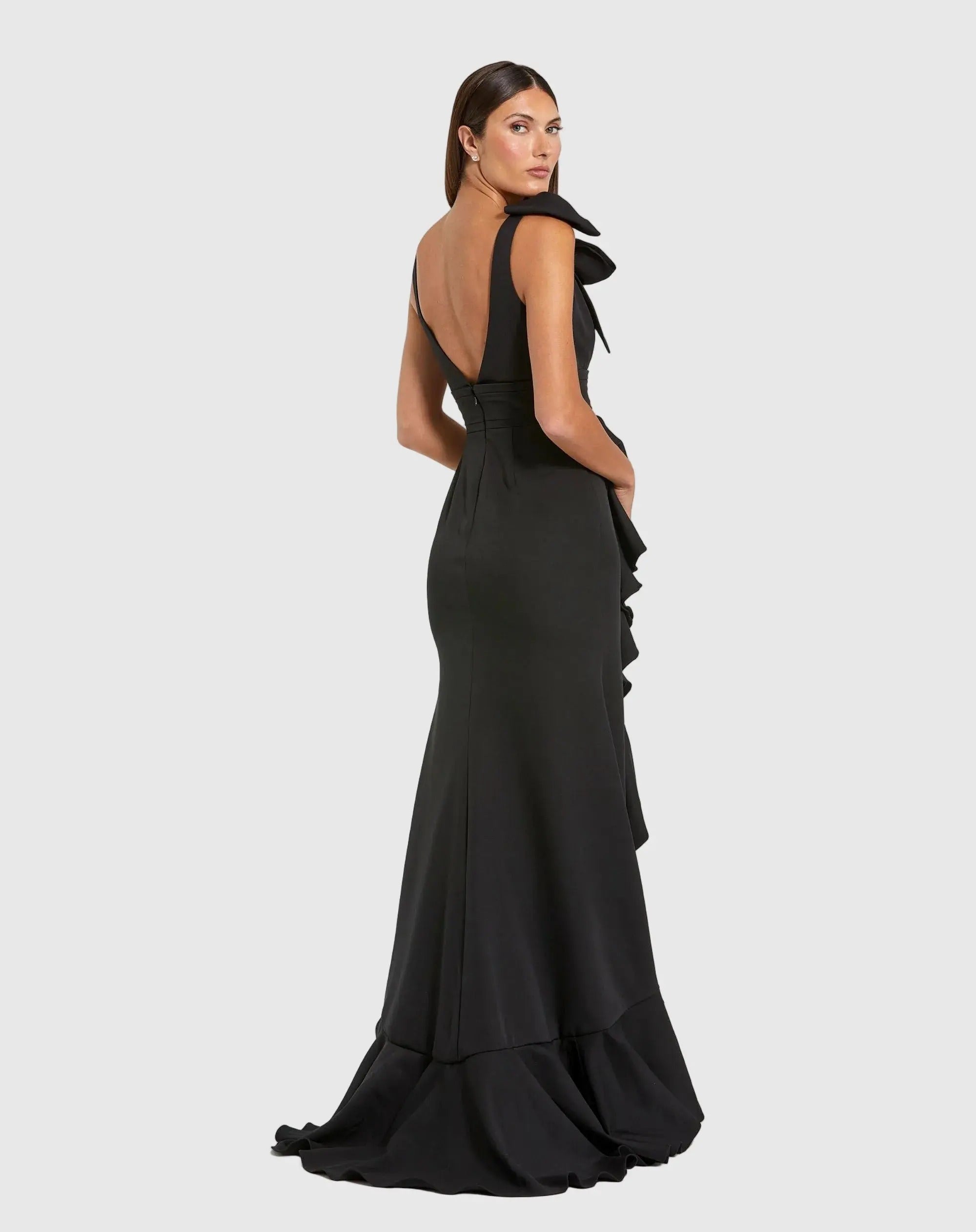 A woman stands against a plain light gray background, wearing the Long Formal Dress: Mac Duggal 49965—a black, floor-length, sleeveless formal gown featuring ruffle details and an open back.